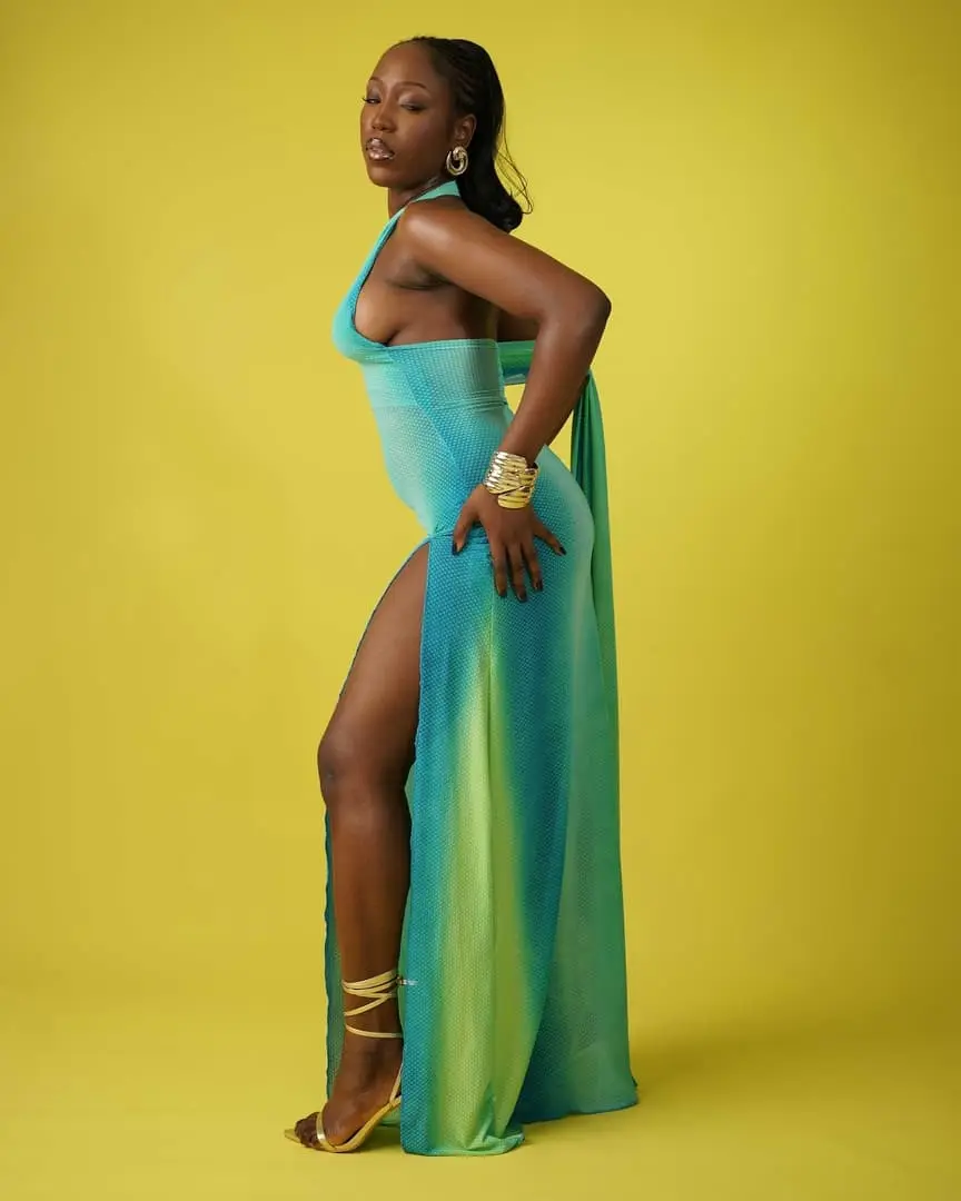 Model in a thigh-slit turquoise-green gradient gown poses in profile against a yellow backdrop.