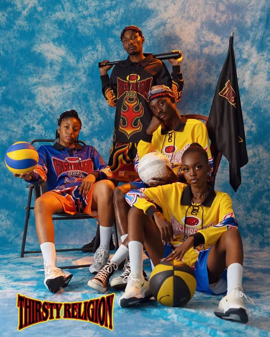 Four models in flame-print Thirsty Religion sports jerseys pose with balls and a bat before a mottled blue backdrop.